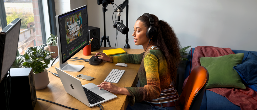 O femeie acasă lucrează la birou cu un MacBook Pro conectat la două monitoare externe, purtând căști și vorbind într-un microfon