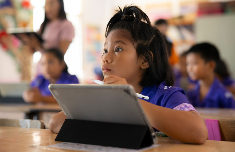 Seorang anak perempuan duduk di mejanya, menatap ke depan kelas dengan penuh antusias. Ia sedang menggunakan iPad dan memegang Apple Pencil.
