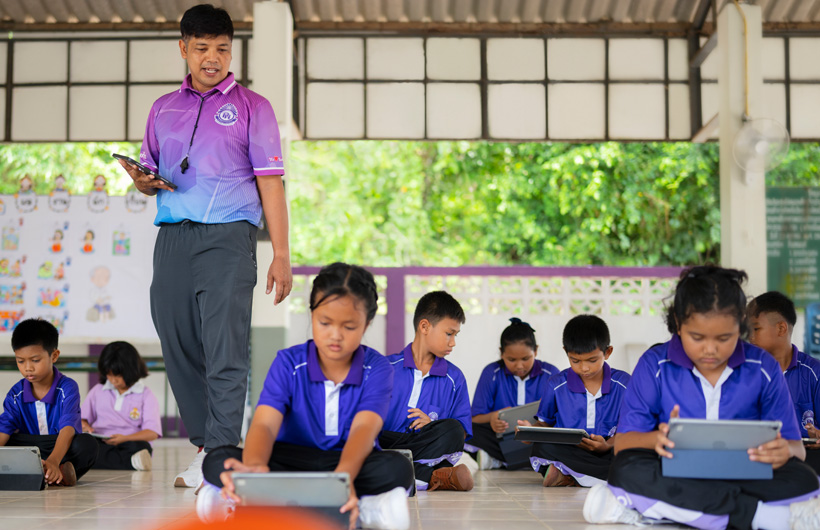 Dalam suasana belajar di alam terbuka, seorang guru pendidikan jasmani berjalan di antara siswanya yang duduk di bawah sambil menggunakan iPad.