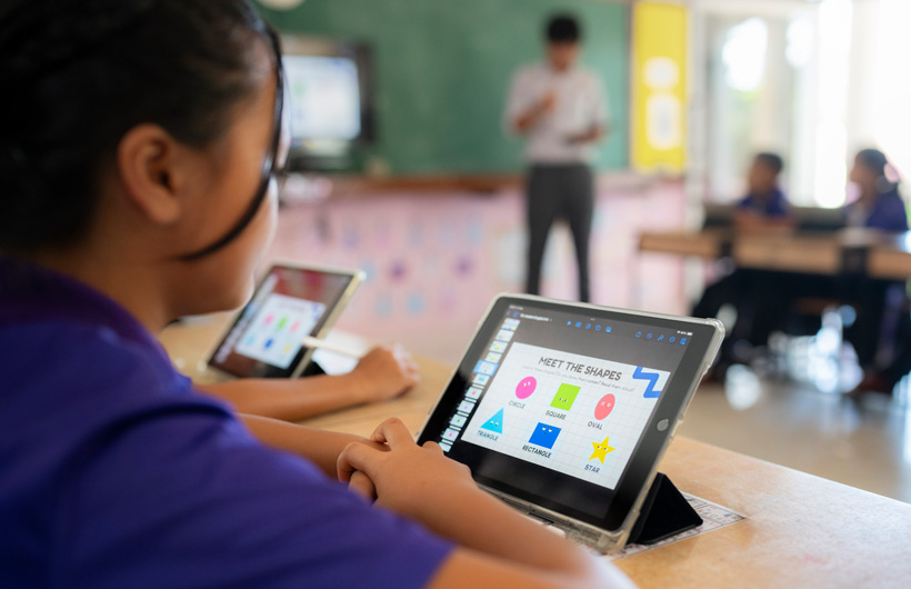 Foto close-up seorang siswa di meja, sedang menggunakan iPad. Di layar, terlihat berbagai bentuk warna-warni dengan judul, 