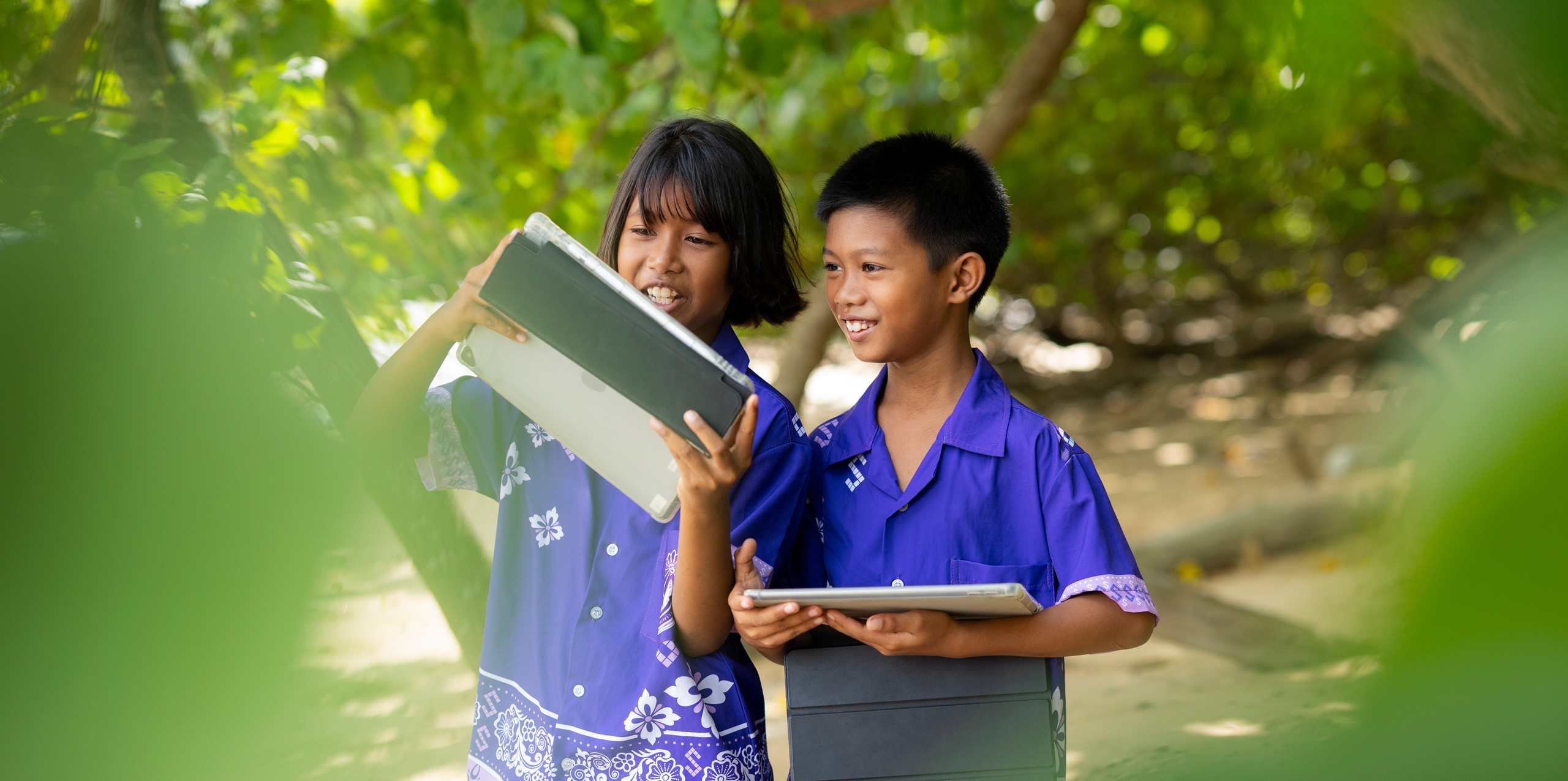 Dua siswa sekolah dasar, satu laki-laki dan satu perempuan, berdiri di luar ruangan, di antara pepohonan tropis, masing-masing memegang iPad. Keduanya melihat layar iPad gadis tersebut, tersenyum, dan tampak fokus.
