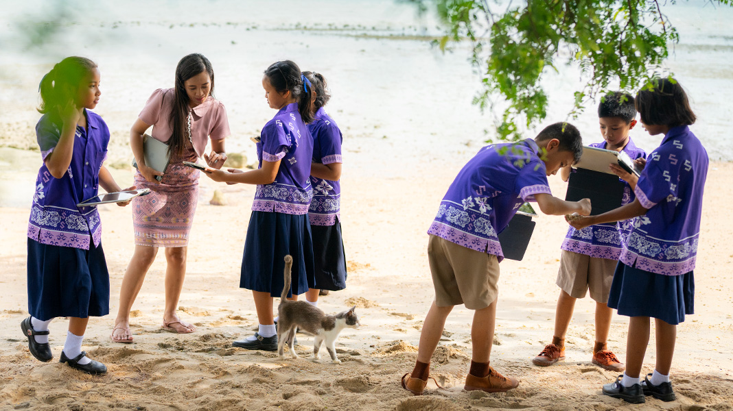 Enam siswa SD dengan seragam ungu cerah berdiri di pantai berpasir sambil memegang iPad. Seorang guru sedang membantu seorang siswa menggunakan iPad, sementara seekor kucing berjalan di sela-sela mereka.